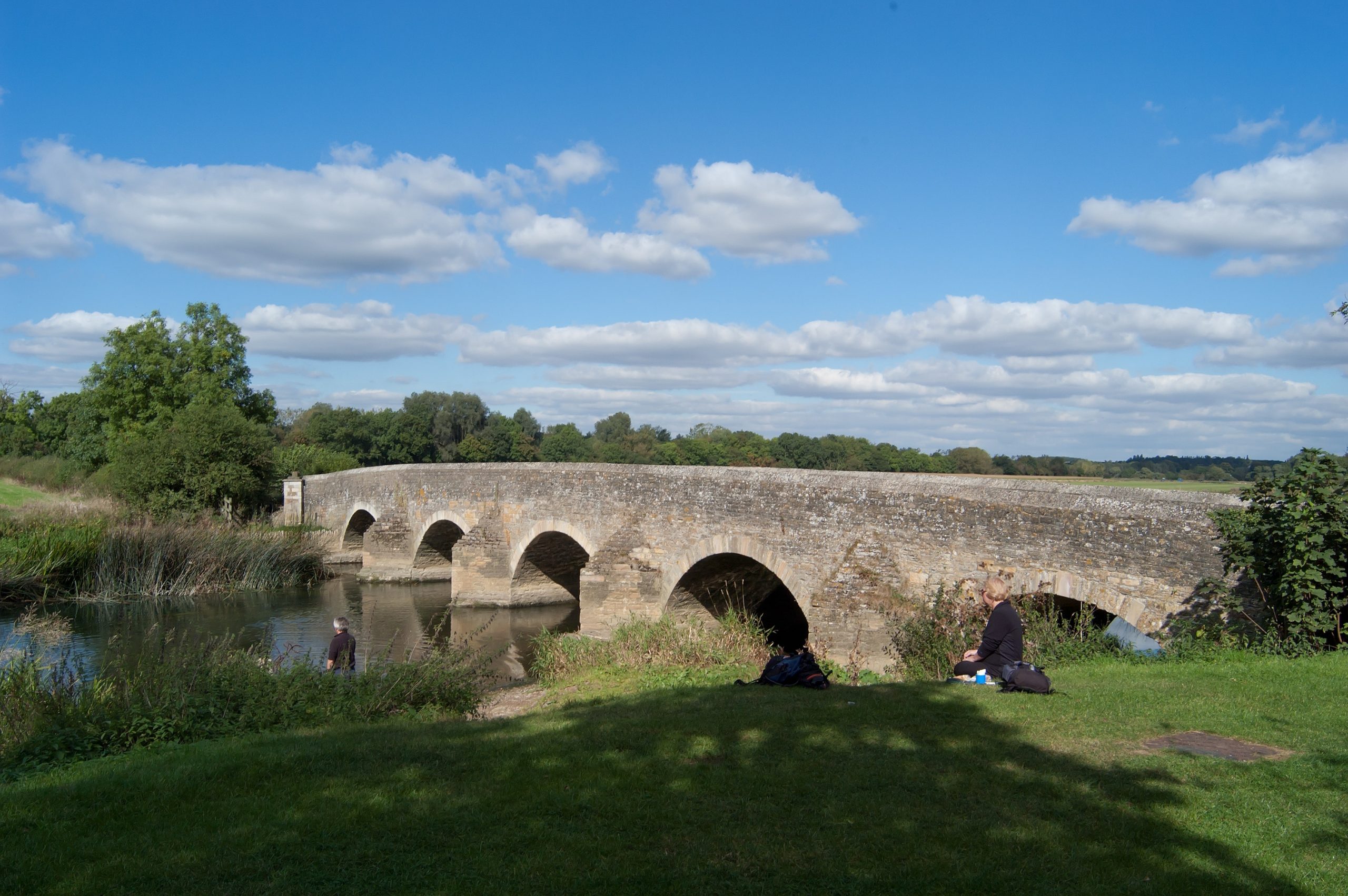 People sitting on a river bank, near a stone bridge