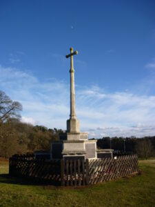 A slender cross on a stone base. The monument is surrounded by a wooden fence.