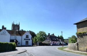 Village shot, church tower rising above a pub and other historic buildings, some of which are thatched.