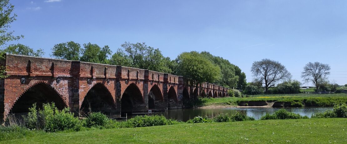 A red brick multi arch bridge over a peaceful river and meadow scene.