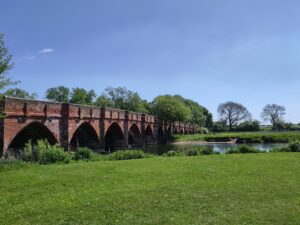 A red brick multi arch bridge over a peaceful river and meadow scene.