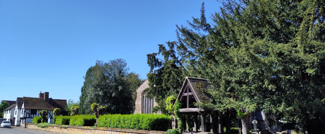 Street scene with low wall, green churchyard with church peeping through and historic houses in the background.