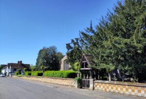 Street scene with low wall, green churchyard with church peeping through and historic houses in the background.