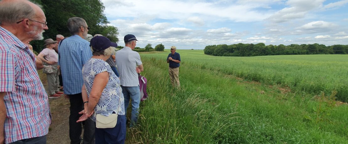 Group of people listening to a man giving a talk. They are stood on the field edge of farmland.