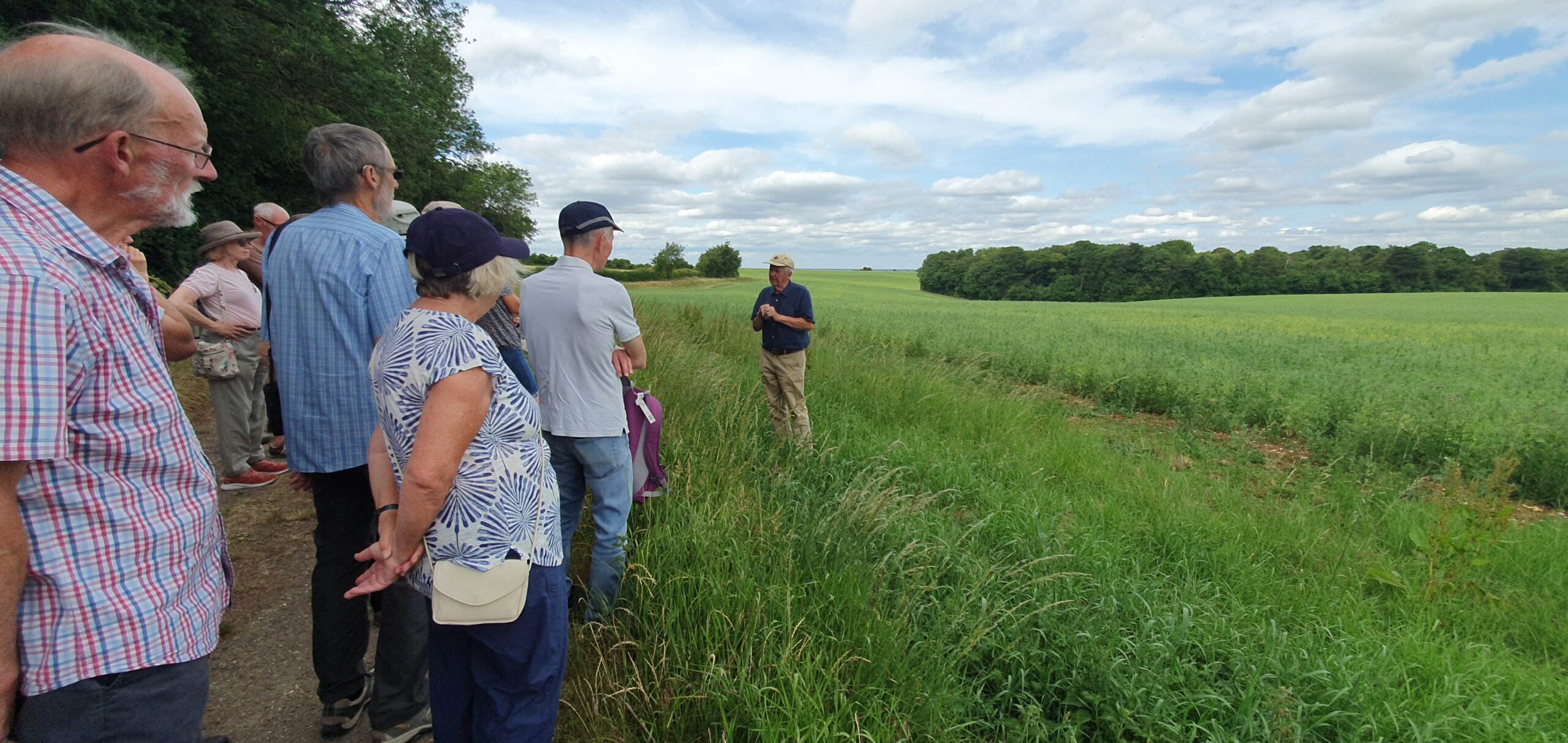 Group of people listening to a man giving a talk. They are stood on the field edge of farmland.