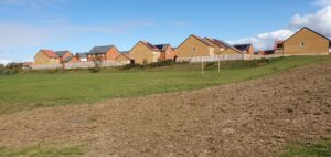 A patch of bare earth with grass and houses behind.
